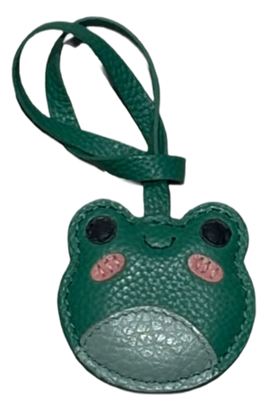 A green faux leather frog-shaped luggage tag with a matching strap, featuring embroidered details for eyes, a smile, and pink cheeks on a white background.