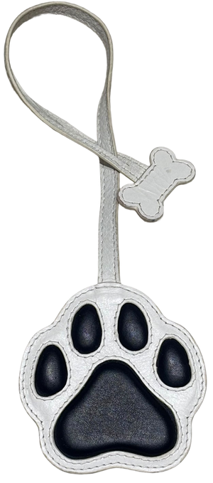 A dog paw print luggage tag or keychain charm with a silver faux leather strap and a matching bone-shaped tag, featuring a black paw pad design on a white background.